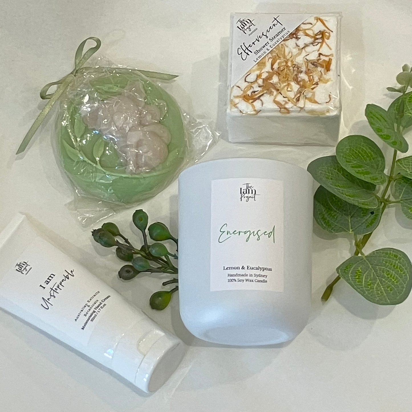 Cosmetic products including a candle, soap, and lotion with greenery on a white surface