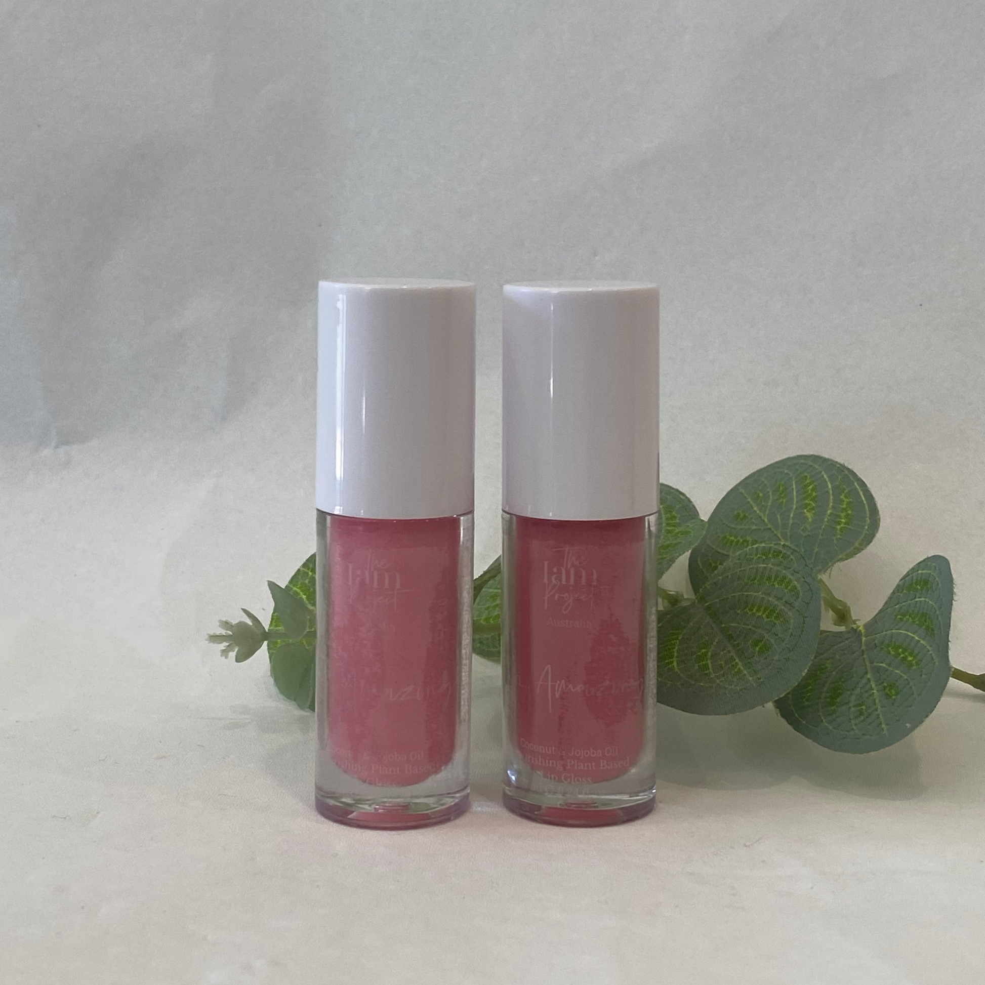 Two pink lip glosses with white caps on a light gray background with green leaves.