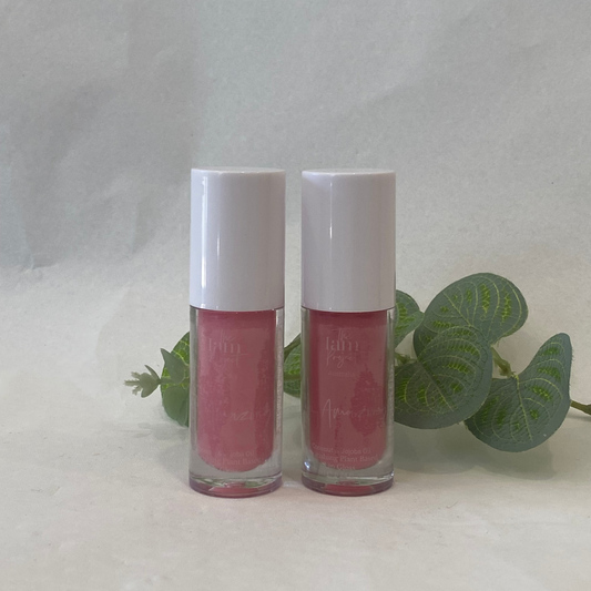 Two pink lip glosses with white caps on a light gray background with green leaves.