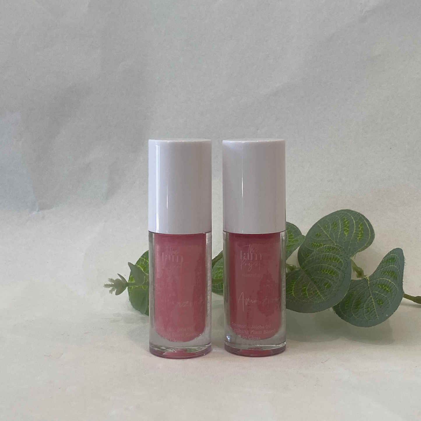 Two pink lip glosses with white caps on a light gray background with green leaves.