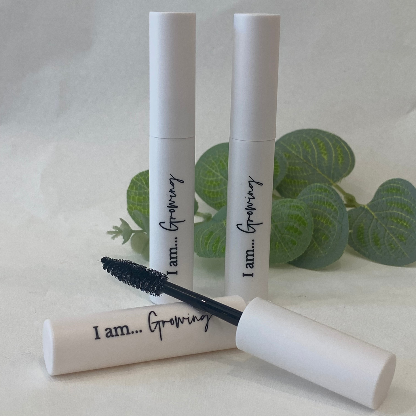 Three 'I am... Growing' eyelash serum tubes on a white background with green leaves.
