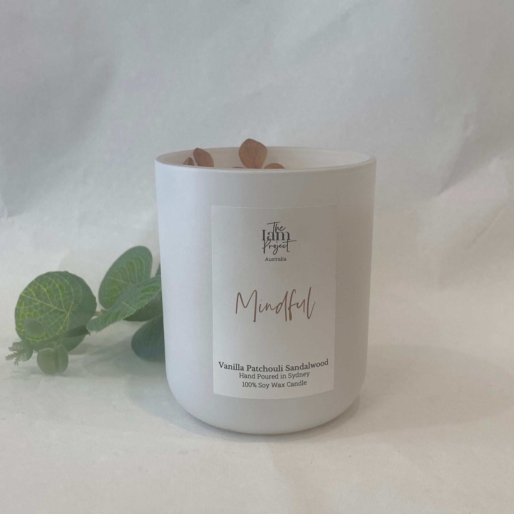 White candle in a glass jar labeled 'Mindful' with eucalyptus leaves on a white background